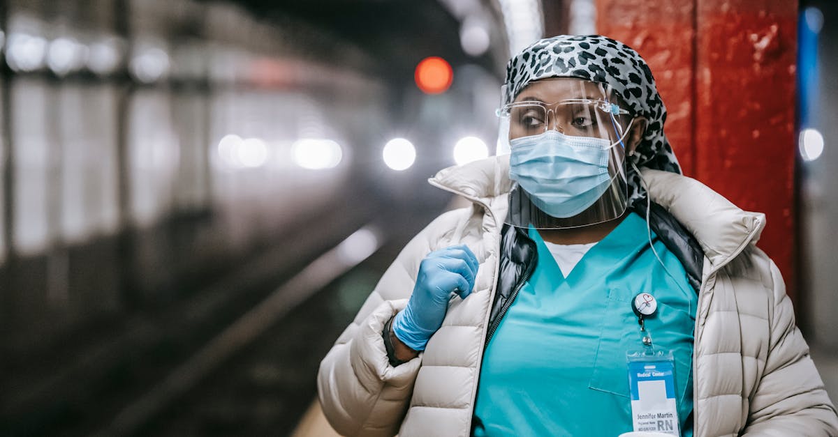 Serious plump African American nurse in uniform and outerwear wearing face mask and latex gloves standing with takeaway coffee standing on metro station platform and looking away thoughtfully